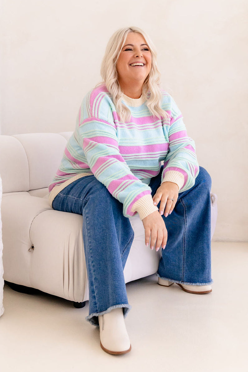 Woman sitting on a white couch wearing a colorful striped sweater and blue jeans.