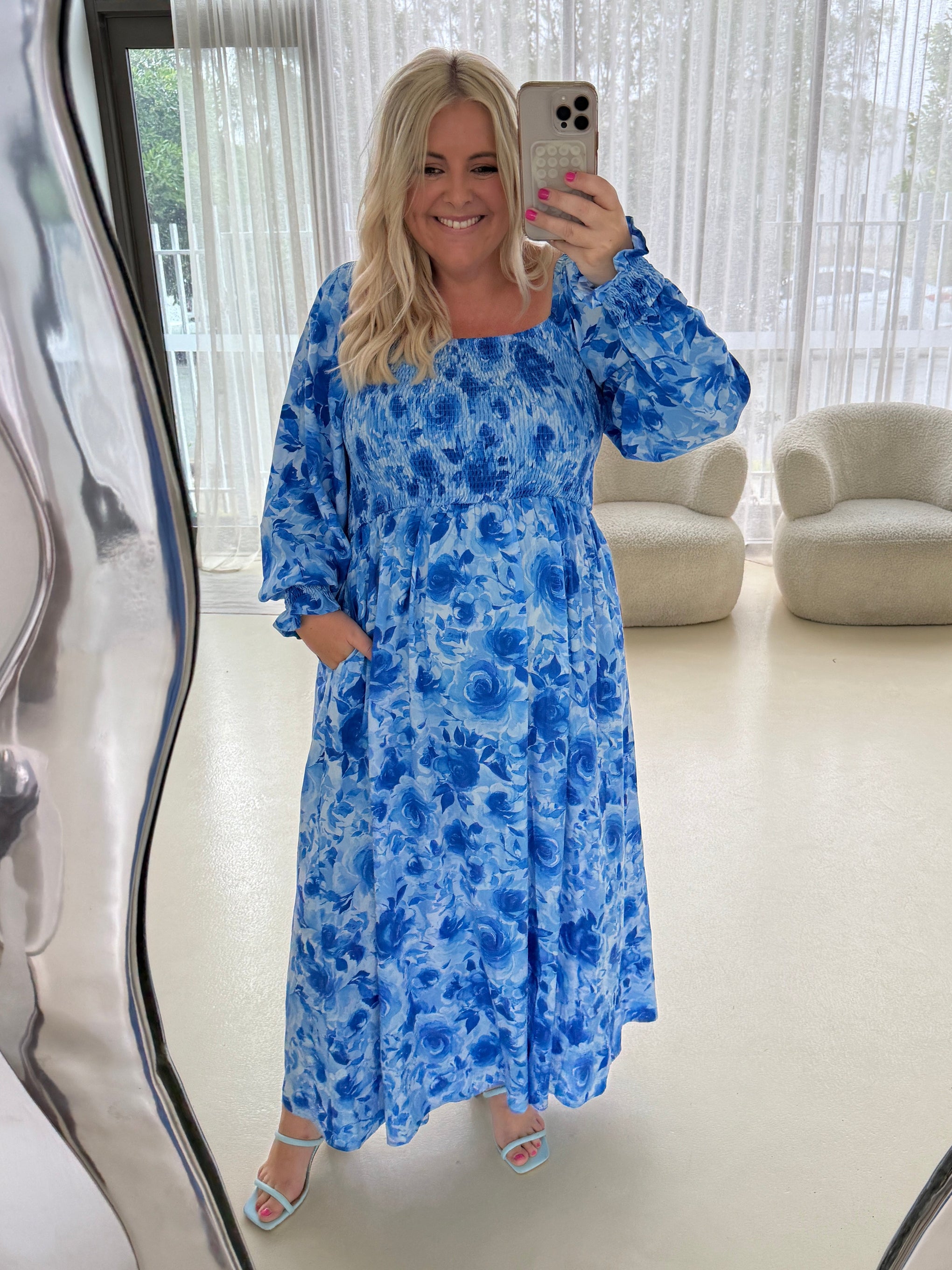 Woman taking a mirror selfie wearing a blue floral dress in a room with a couch and window blinds.